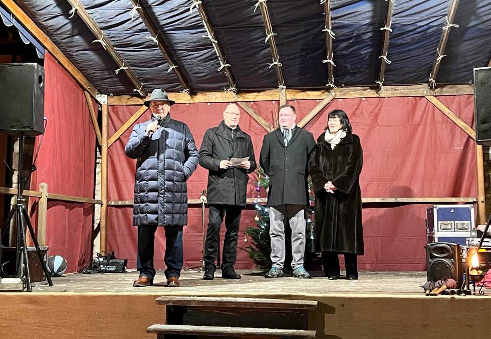 Drei Männer und eine Frau stehen am Abend, es ist dunkel, dick angezogen, weil Winter auf einer Bühne. Sie schauen alle nach vorne. Der Ministerpräsident hat das Mikro in der Hand und redet.