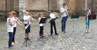 7 Kinder stehen mit Trompeten und Posaunen und musizieren. Vor ihnen sind Notenständer.