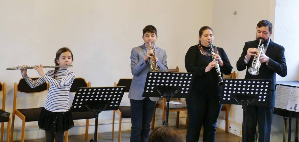 von links nach rechts stehen: ein Mädchen mir einer Querflöte, ein ältere Junge mit einer Trompete, eine Frau mit Klarinette und eine Mann mit Trompete. Vor ihnen stehen schwarze Notenständer.