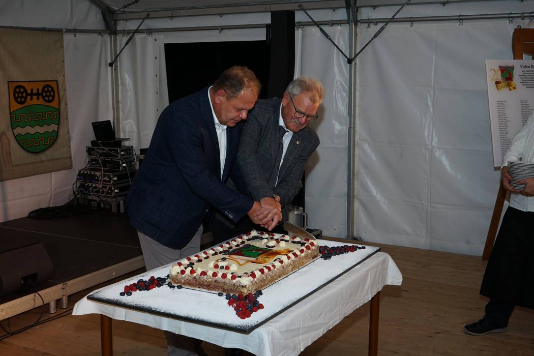 Auf einem Tisch ist eine viereckige Geburtstagstorte auf der 20 Jahre Alte Brennerei steht. Herr Prinzing und Herr Kupski schneiden die Torte an.