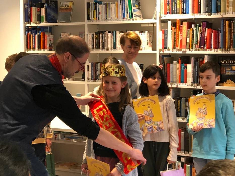 Ein Bücherregal mit vielen Büchern im Hintergrund. Davor stehen ein Mädchen und ein Junge und halten ihre Urkunden in den Händen. Ganz vor wird Elsa Klingner eine rote Schärpe überreicht. Sie trägt eine Krone und lacht in die Kamera. Ein Mann hängt ihr die Schärpe um.