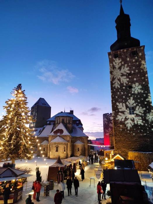 Die Querfurter Burganlage, rechts ein Turm, der mit Schneekristallen angestrahlt wird, daneben die Kirche und links ein beleuchteter Weihnachtsbaum. Dazwischen Buden und Stände. Es sind Menschen unterwegs. Alles ist verschneit. Es wird langsam dunkel.