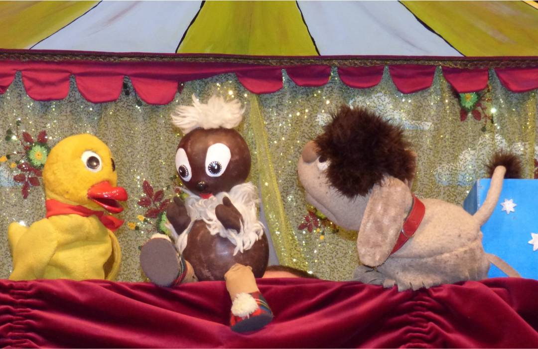 Die Puppen: das gelbe Schnatterinchen. der braune Kobold Pittiplatsch und der braune Hund Moppi sitzen auf einer Puppentheaterbühne. Diese ist rot.