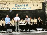 Paul Wanzek, Hartmut Handschak und Torsten Ringling stehen auf der Bühne. Hinter ihnen steht ein Chor über dem ein Banner mit Chorfest  des Saalekreises hängt.