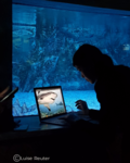 Es ist dunkel. Vor einem beleuchteten großen Aquariumfenster sitzt ein Mädchen mit einem Laptop. Auf dem Bildschirm ist ein Hai zu sehen.