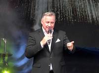 Ein Mann im Anzug, der singt. Er ht ein Mikrofon in der rechten Hand, die linke zeigt mit dem Zeigefinger nach vor.